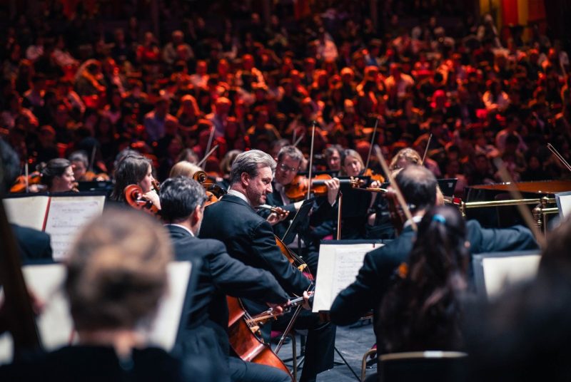 The Royal Philharmonic Orchestra performs on stage in front of a large, warmly lit audience. Musicians in formal black attire play string instruments, with sheet music visible on stands. The focus is on a cellist at the center, with other players surrounding him in close formation. The crowd in the background appears engaged, creating a vibrant, immersive atmosphere.