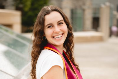 Young woman with long dark hair, wearing a white dress and graduation stole, sitting on a stone wall