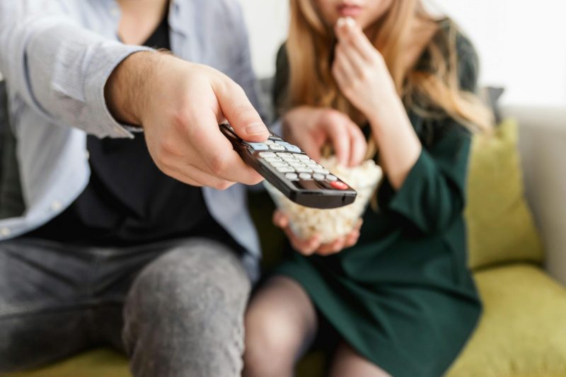 A man and a woman, sitting on a couch, eat popcorn and point a remote toward an offscreen television. 