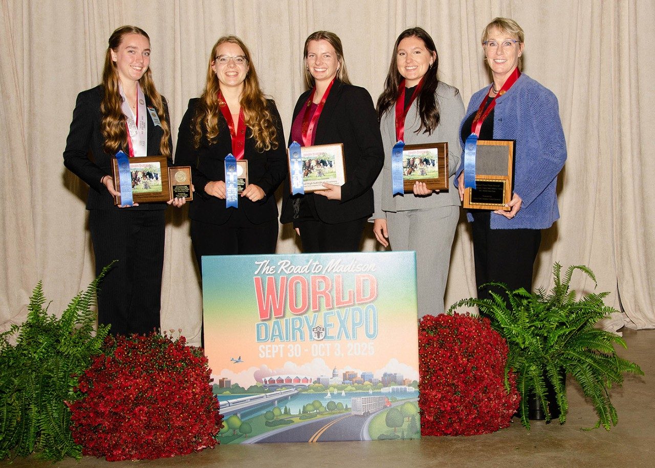 Five women stand indoors in front of a beige curtain, smiling and holding plaques and blue ribbons. They are dressed in professional attire and wearing red medals around their necks. In front of them is a colorful sign reading “The Road to Madison – World Dairy Expo, Sept 30 – Oct 3, 2025,” surrounded by green ferns and red flowers.