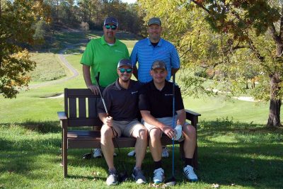 Team Farm Credit Maroon. Two team members sitting on a bench, the other two behind the bench in the shade of a tree.