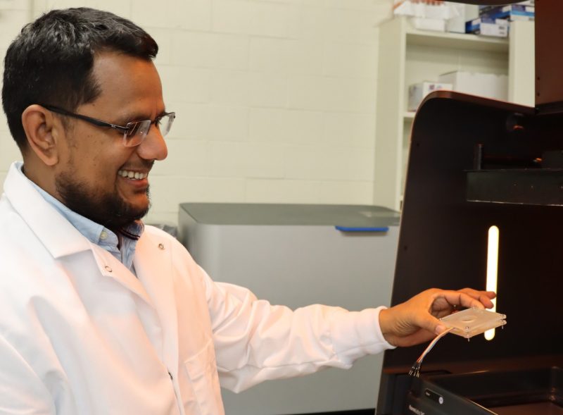 Smiling researcher wearing lab coat and holding a 3-D printed biosensor.