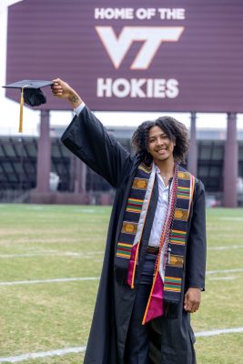 Brandon Duncan standing in front of Lane Stadium with his graduation gown and stoles on. He is holding his cap in the air.