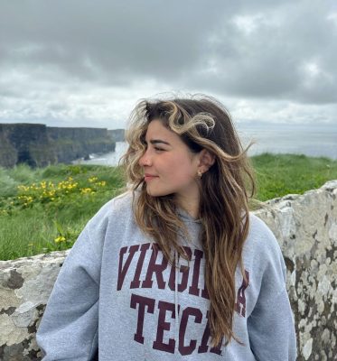 A young woman in a Virginia Tech hoodie looking to the left. She is standing in front of a stone wall overlooking the ocean.
