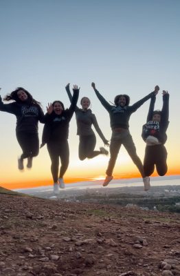 A group of young adults jumping together on a hill in front of the sunset.