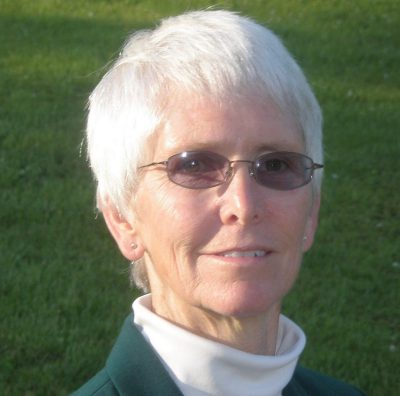 An older woman with short white hair, wearing glasses, a dark green jacket, and a white turtleneck, smiling slightly outdoors on a sunny day with grass in the background.