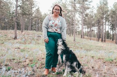 Megan Arant standing with her dog in front of tall pines.