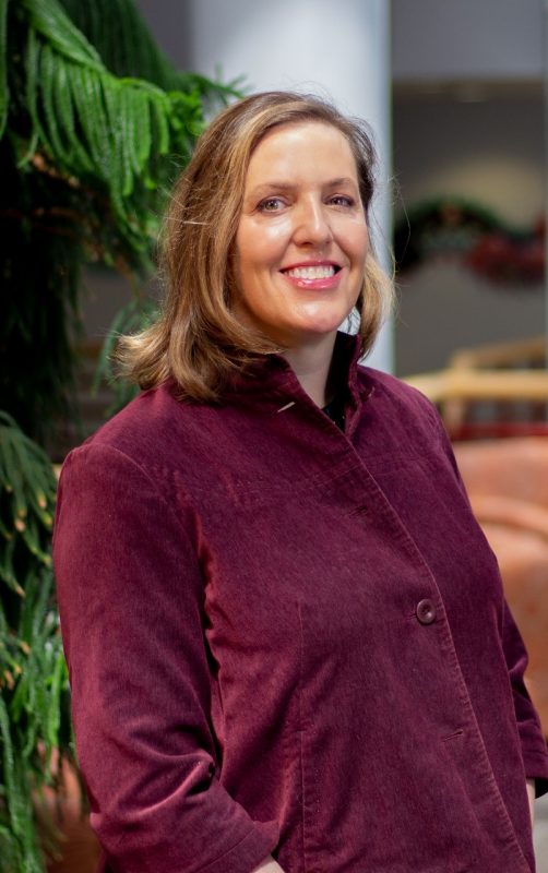 Dr. Erica Feurerbacher at the Holtzman Alumni Center. Evergreen and white columns in the background. Cropped vertically.