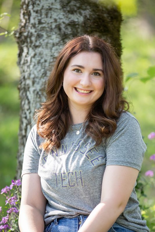 Smiling young woman sitting in front of a tree with purple flowers behind her.