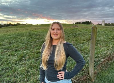 Young woman with long blond hair standing in front of a fenced field at sunset. Clouds, trees, and a silo in the background.