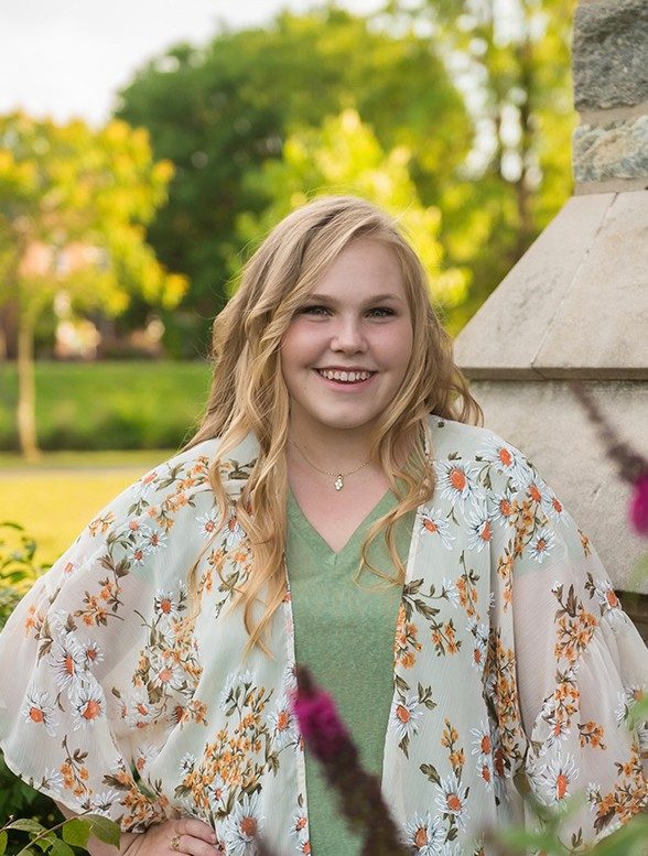 Young woman with long blond hair standing by a stone column and smiling. Green trees and grass in the background.