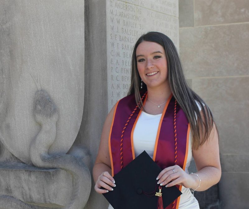 Young woman with lon dark hair standing at the Pylons on Virginia Tech campus wearing graduation cord, stole, and holding graduation cap with a 2025 tassell.