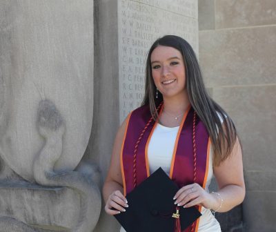 Young woman with lon dark hair standing at the Pylons on Virginia Tech campus wearing graduation cord, stole, and holding graduation cap with a 2025 tassell.