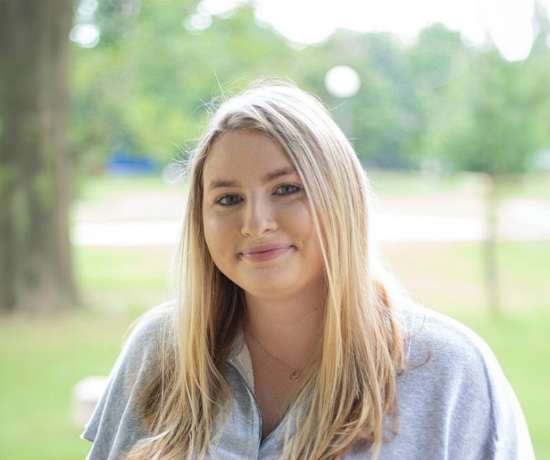 Smiling blond woman with a blurred background of greenery and trees.