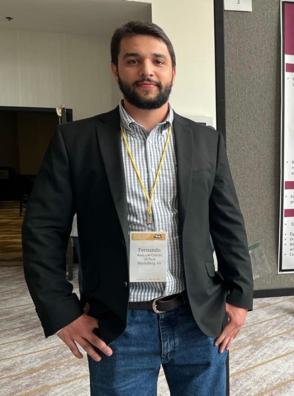 Young man with dark hair and beard wearing a blazer, collared shirt, and dark blazer with an identification lanyard at a research symposium.