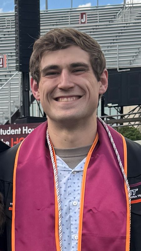 Smiling young man wearing graduation robe, stole, and cord at Lane Stadium. Bleacher section J in the background.