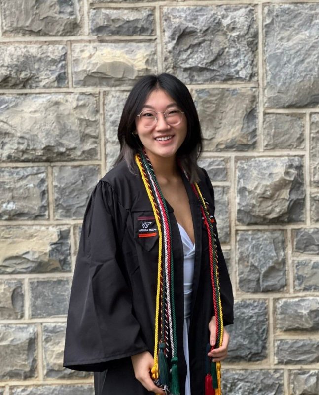 Young woman with dark hair and glasses smiling in her graduation regalia in front of a HokieStone wall. Virginia Tech campus.