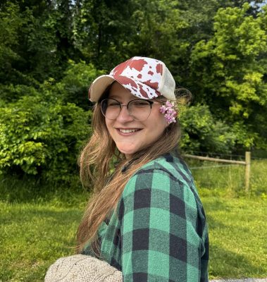 Smiling young woman with long hair and glasses wearing a cap and green flannel shirt. Green trees and grass in the background.