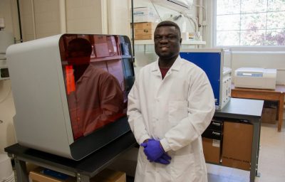 Young man dressed in lab coat and gloves beside laboratory equipment.