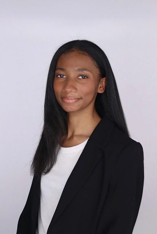 Professional photo of Kennedi Essesex wearing a  white shirt and dark blazer against a pale grey backgound. Yound woman with dark hair looking toward the camera.