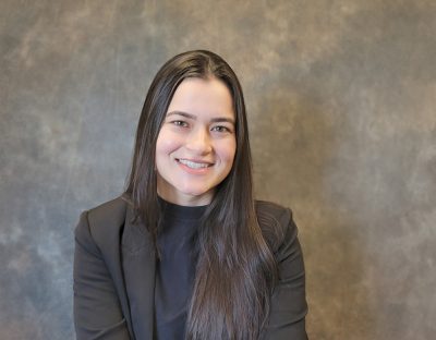 Smiling young woman with long dair hair wearing a black blazer and dark shirt. Mottled gray background.