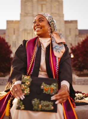 Young black woman wearing graduation regalia, holding a decorated mortarboard cap. Maroon foliage and Burruss Hall in the background.