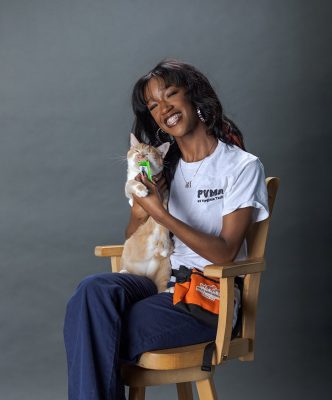 Young woman smiling while holding a cat, seated in a wooden chair in front of a gray backdrop.