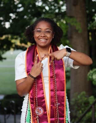 Smiling graduate wearing glasses, a white dress, and maroon and orange honor stoles with cords, standing outdoors and making a hand gesture in the shape 'V'T with her fingers.