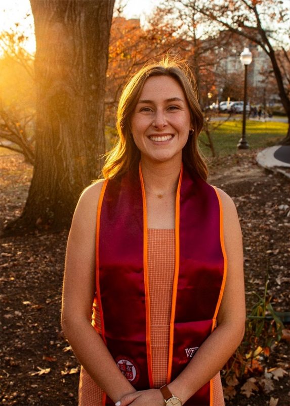 White woman with shoulder-length brown hair smiling, standing in front of a tree in late autumn with leaves on the ground. Buildings in the background.