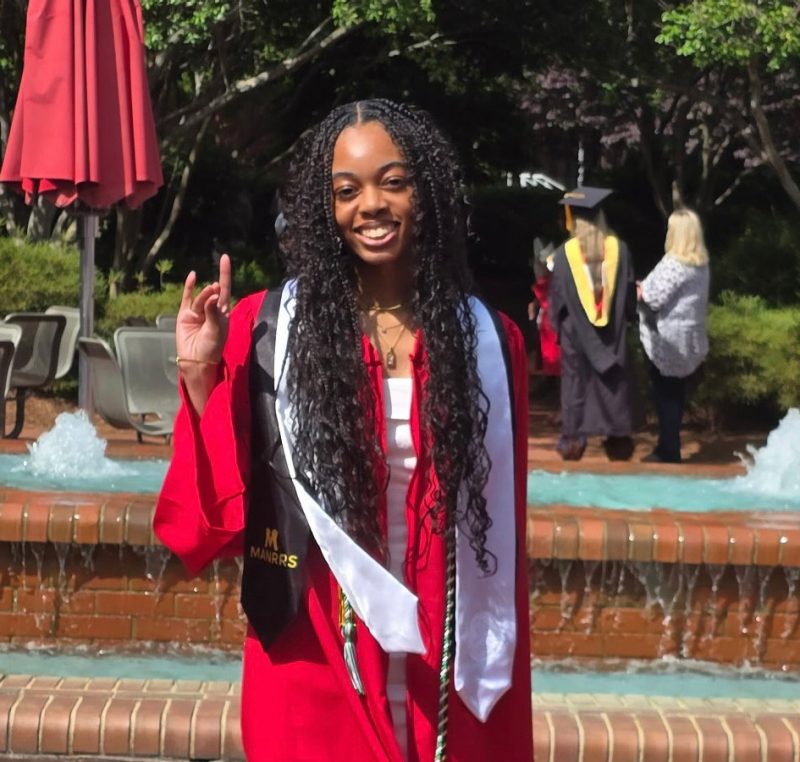 Smiling graduate wearing a red gown, white and black stoles, and honor cords, standing in front of a brick fountain with water flowing, holding up a hand gesture with two fingers raised.