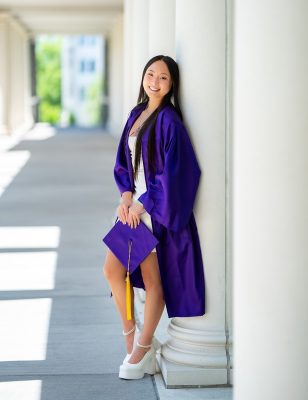 Young woman with long dark hair, wearing a purple graduation gown, holding a matching mortar with a gold tassel, leaning against a white column in an open portico.