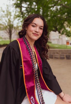 Smiling young woman with dark hair wearing a white dress under her graduation  gown, stole, and multiple cords. Trees and a HokieStone retaining wall in the background.