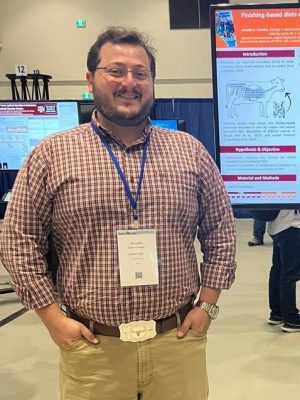 Young man with brown hair and short beard wearing a maroon plaid shirt and dunn pants standing in front of his poster presentation.