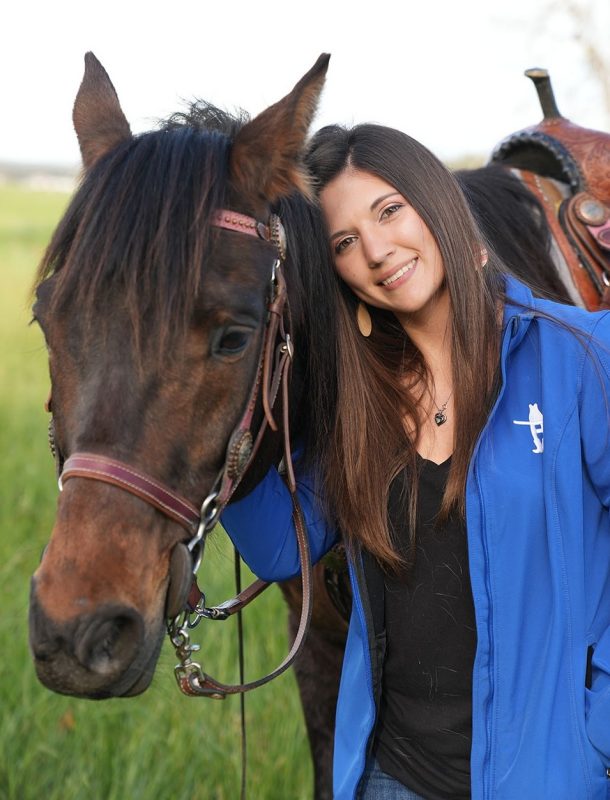 Smiling young woman with long brown hair leaning into a horse with a bridle and saddle.