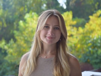 A young woman with long blonde hair smiles softly while standing outdoors, with green and yellow foliage blurred in the background.