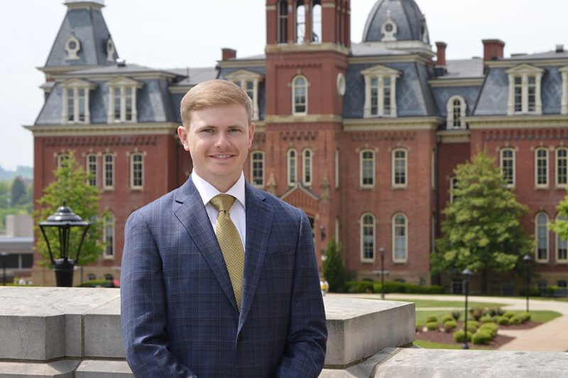 Young man with sandy hair dressed in a suit and tie standing in front of an impressive brick structure with bell tower, mansard roofline, chimneys, and distintive architecture.