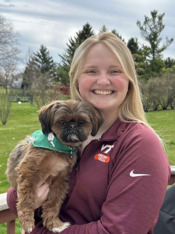 Young woman with long blond hair holding a small dog wearing a green bandana decorated with white ducks wearing bowties.