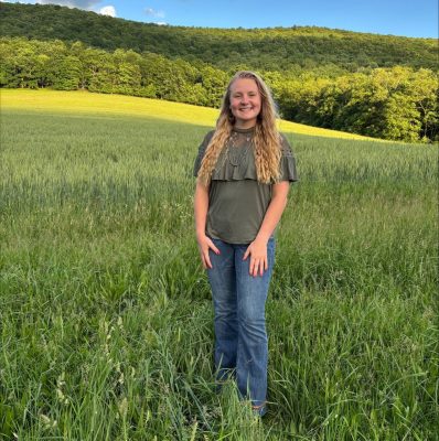 Smiling young woman in blue jeans standing in a lush green pasture with green forest in the far background, under a bright blue sky dotted with puffy white clouds.