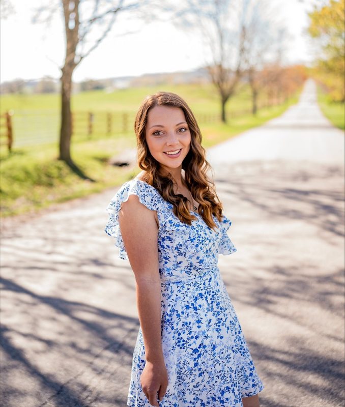 Smiling young woman in a white and blue floral dress standing on a straight road, facing camera. Pasture and bare trees in the background.