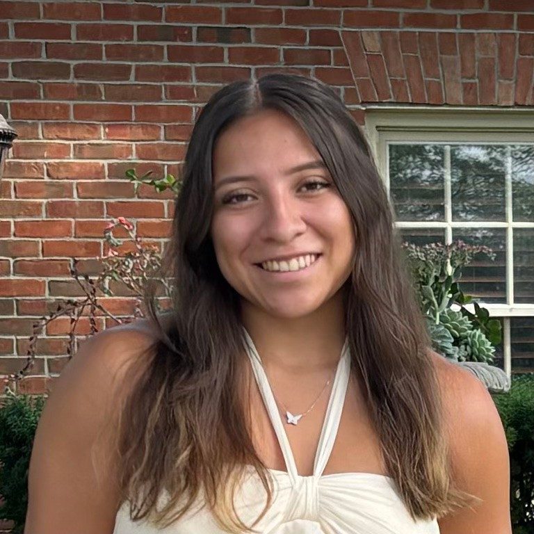 Smiling young woman with long dark hair in front of a brick exterior wall with garden plants and a window.