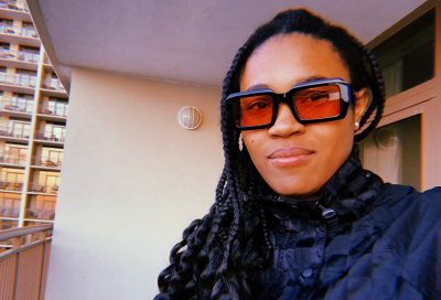 Young woman with long braids and glasses standing on a balcony, looking at the camera.