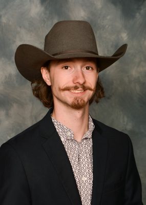 Professional photo of a smiling young man wearing a cowboy had and dark blazer against mottled blue-gray backdrop.