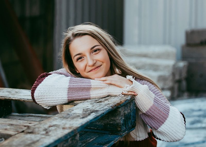 Smiling young woman with long brown hair leaning on a wooden farm wagon.