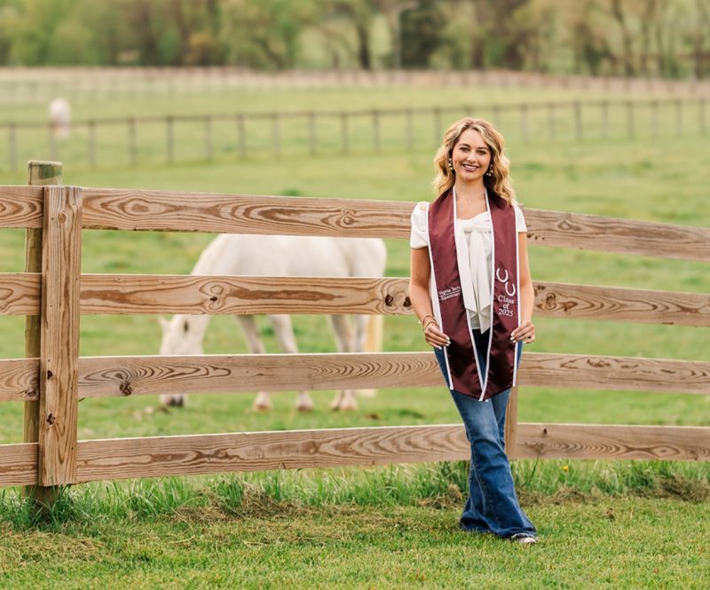 Young woman with shoulder length blond hair wearing a maroon Virginia Tech Class of 2025 Equestrian graduation stole with jeans and a white shirt, standing in front of wooden fence with a white horse.