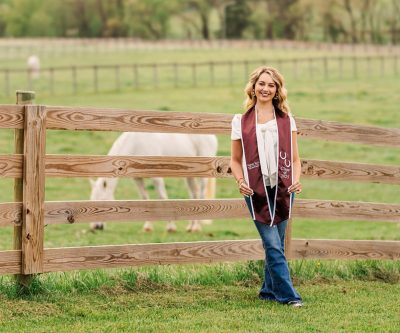 Young woman with shoulder length blond hair wearing a maroon Virginia Tech Class of 2025 Equestrian graduation stole with jeans and a white shirt, standing in front of wooden fence with a white horse.
