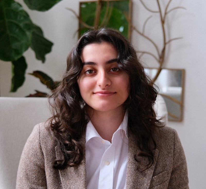 Young woman with long dark hair wearing a herringbone blazer and white shirt.