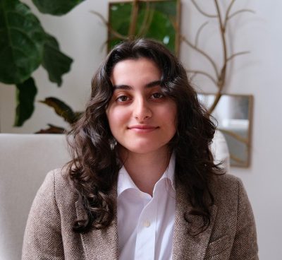 Young woman with long dark hair wearing a herringbone blazer and white shirt.