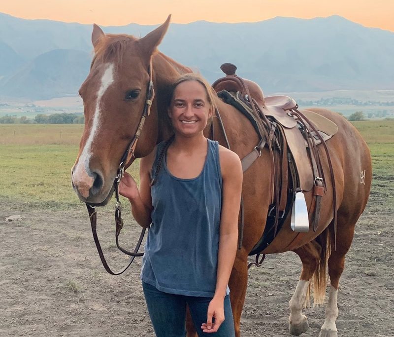 Young woman with dark hair pulled into a ponytail standing with a saddled horse smiling. Mountains in the background.