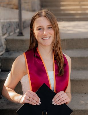 Young woman wih long reddish brown hair smiling, sitting on HokieStone steps with her graduation cap.