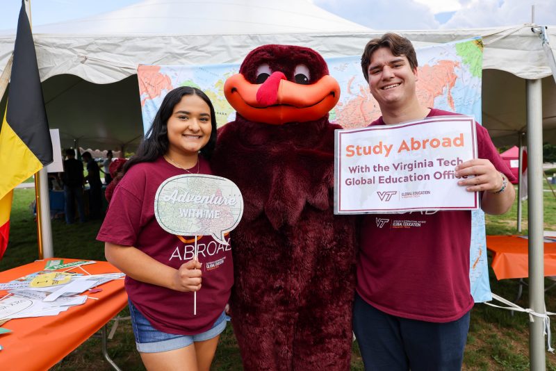 The Hokie Bird is flanked by two students holding signs promoting study abroad